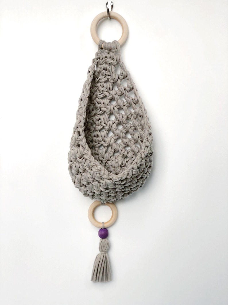 May include: A gray crocheted hanging basket with a wooden ring at the top and a wooden ring with a purple bead and tassel at the bottom.