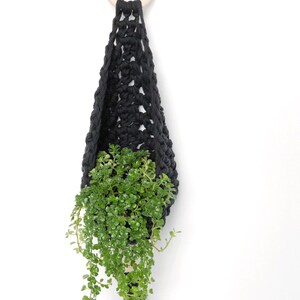 May include: A black macrame plant hanger with a wooden ring at the top. A green plant is hanging in the macrame hanger.