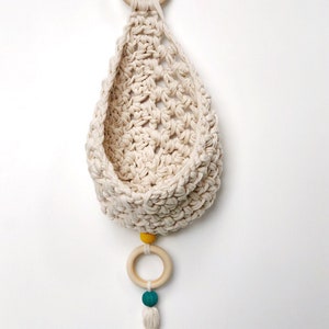 May include: A white crocheted hanging basket with a wooden ring and yellow and teal beads. The basket has a tassel at the bottom.