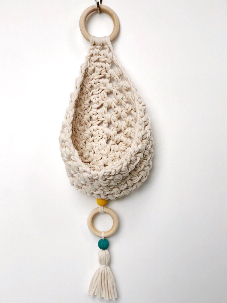 May include: A white crocheted hanging basket with a wooden ring and yellow and teal beads. The basket has a tassel at the bottom.