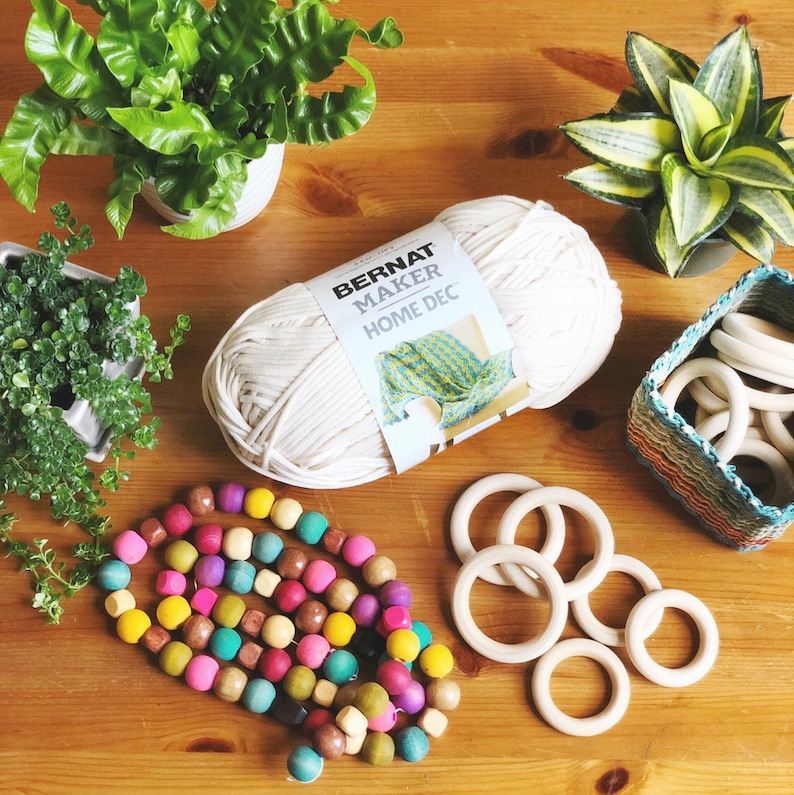 May include: A ball of white Bernat Maker Home Dec yarn, a wooden bead garland with colorful beads, and a basket of wooden rings.
