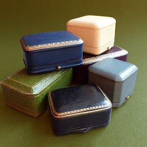 May include: A collection of six vintage jewelry boxes in various shapes and colors, including a blue box with gold trim, a green textured box, a cream-colored box, a dark red box, a black box with gold trim, and a grey box with a hinged lid.