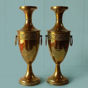 Peut inclure: Une paire d'urnes en laiton poli dotées d'un col fin, d'un bord évasé et d'un corps arrondi. Chaque urne est ornée d'une bande gaufrée décorative à motifs de feuilles et comprend deux poignées circulaires montées sur les côtés supérieurs.