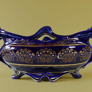 May include: A cobalt blue ceramic bowl with gold floral designs and trim. The bowl has a unique shape with handles and rests on three small feet. The gold detailing includes a band of floral patterns and accents along the edges.