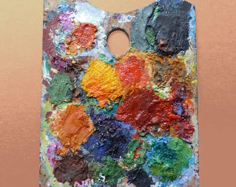 May include: A rectangular wooden artist palette featuring a thumb hole at the top center. The surface is covered in thick, textured dabs of oil paint in various colors including yellow, orange, red, green, blue, brown, black, and white.