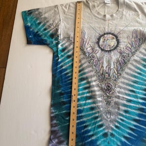 Puede incluir: Una camiseta tie-dye con un esquema de color azul, blanco y gris. La camiseta presenta un gr&aacute;fico de una luna rodeada de cristales y un dise&ntilde;o geom&eacute;trico.