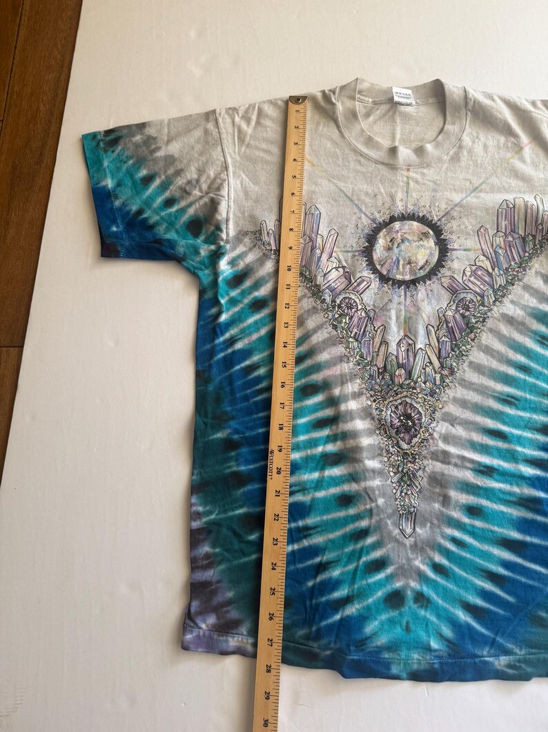 Puede incluir: Una camiseta tie-dye con un esquema de color azul, blanco y gris. La camiseta presenta un gr&aacute;fico de una luna rodeada de cristales y un dise&ntilde;o geom&eacute;trico.