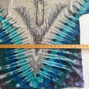 Puede incluir: Una camiseta tie-dye con un dise&ntilde;o de cristal en el centro. La camiseta es de color gris claro con acentos tie-dye azules y morados.