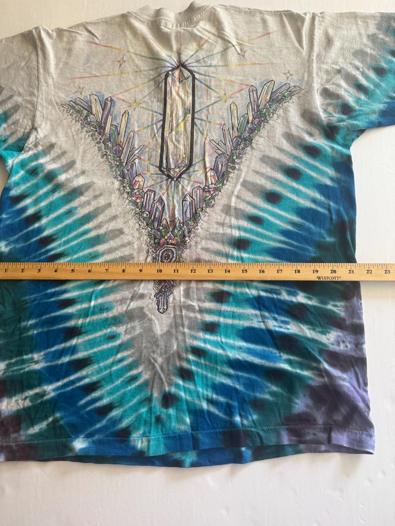 Puede incluir: Una camiseta tie-dye con un dise&ntilde;o de cristal en el centro. La camiseta es de color gris claro con acentos tie-dye azules y morados.