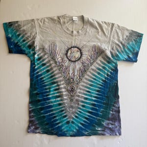 Puede incluir: Una camiseta tie-dye con un patr&oacute;n azul y gris. La camiseta presenta un gr&aacute;fico de una luna rodeada de cristales.