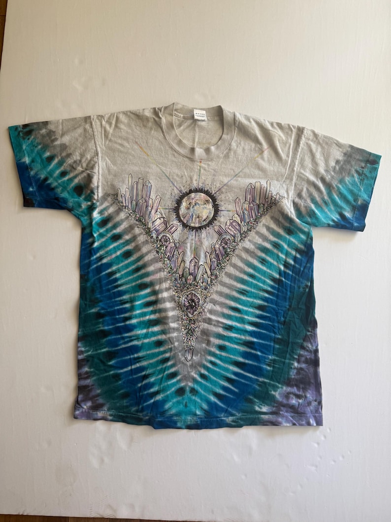 Puede incluir: Una camiseta tie-dye con un patr&oacute;n azul y gris. La camiseta presenta un gr&aacute;fico de una luna rodeada de cristales.