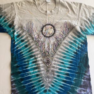 Puede incluir: Una camiseta tie-dye con un esquema de color azul, verde y blanco. La camiseta presenta un gr&aacute;fico de una luna rodeada de cristales.