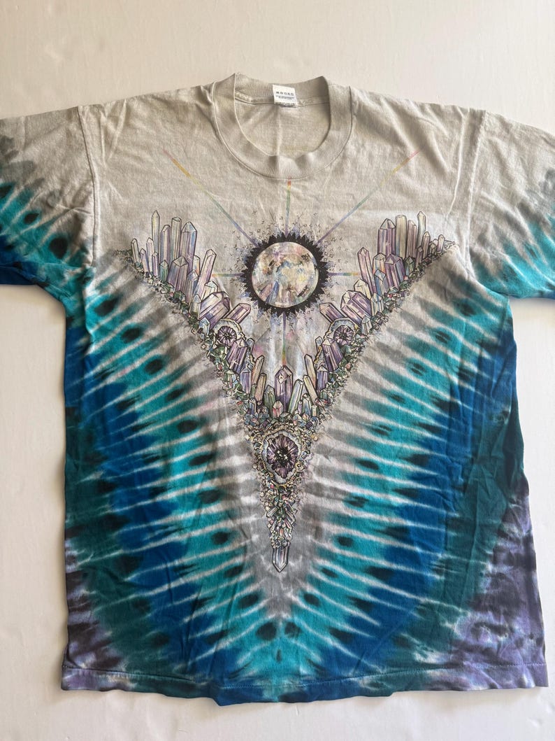 Puede incluir: Una camiseta tie-dye con un esquema de color azul, verde y blanco. La camiseta presenta un gr&aacute;fico de una luna rodeada de cristales.