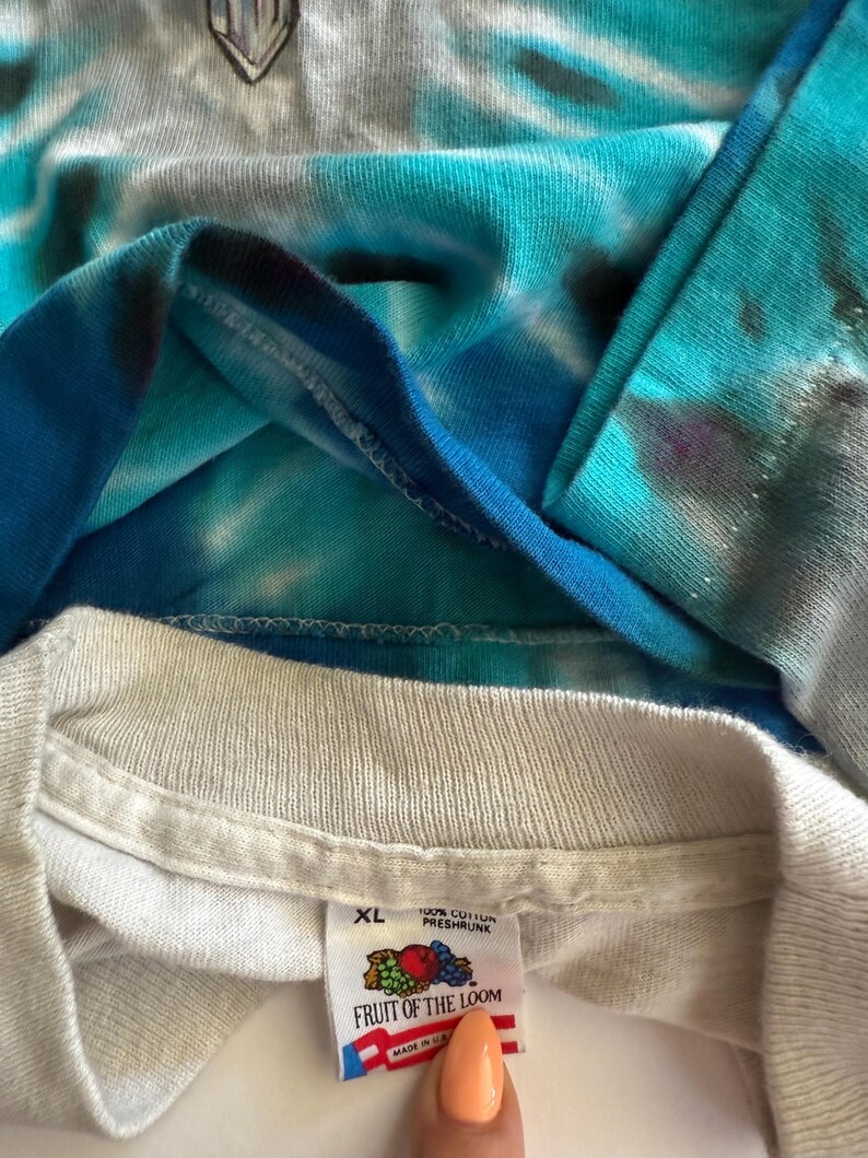 Puede incluir: Una camiseta blanca con un patr&oacute;n de tie-dye azul y blanco. La camiseta tiene una etiqueta de Fruit of the Loom con la talla XL y el texto "100% Algod&oacute;n Preencogido" y "Fruit of the Loom" impreso en ella. La etiqueta tambi&eacute;n dice "Hecho en EE. UU.".