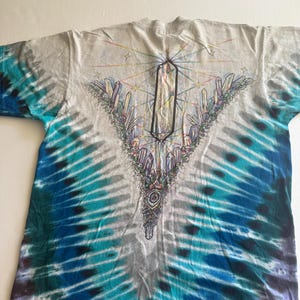 Puede incluir: Una camiseta tie-dye con un esquema de color azul, verde y blanco. La camiseta presenta un dise&ntilde;o de cristal con un cristal grande en el centro y cristales m&aacute;s peque&ntilde;os que lo rodean. El dise&ntilde;o est&aacute; rodeado por un arco&iacute;ris de colores.