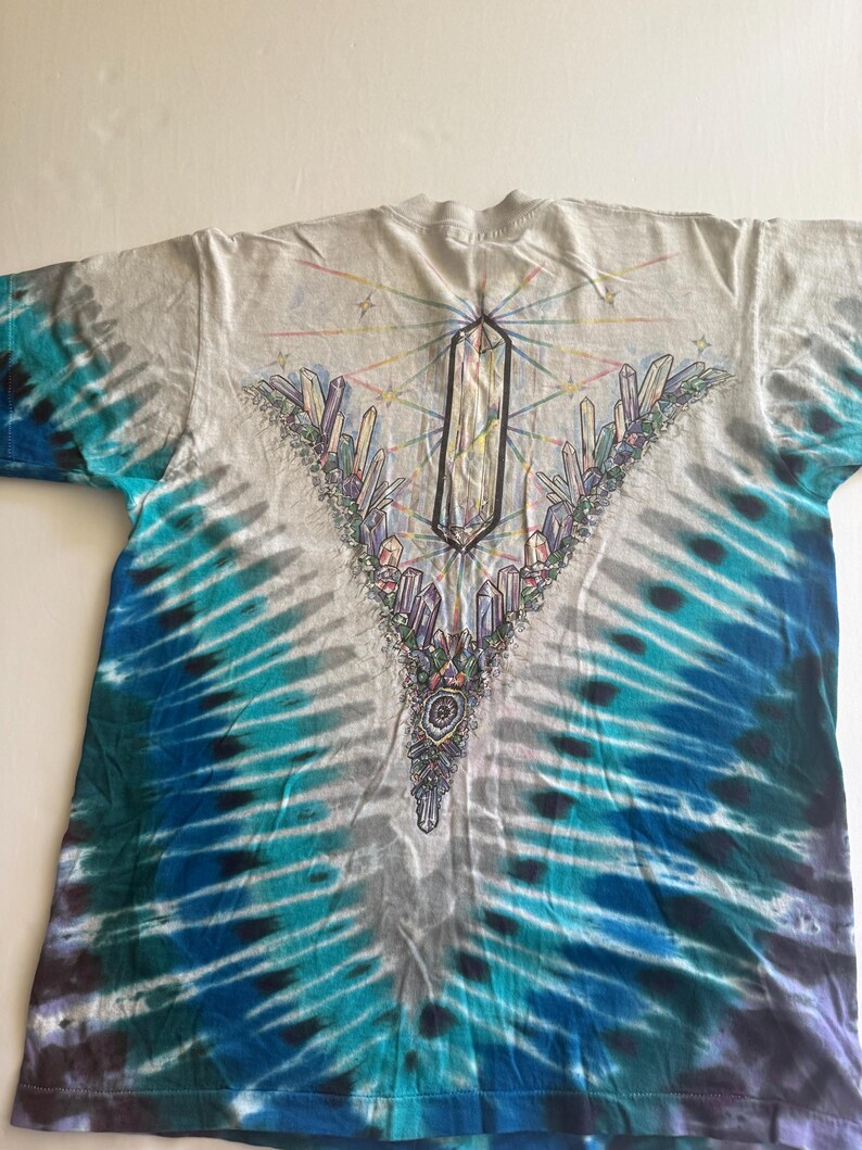 Puede incluir: Una camiseta tie-dye con un esquema de color azul, verde y blanco. La camiseta presenta un dise&ntilde;o de cristal con un cristal grande en el centro y cristales m&aacute;s peque&ntilde;os que lo rodean. El dise&ntilde;o est&aacute; rodeado por un arco&iacute;ris de colores.