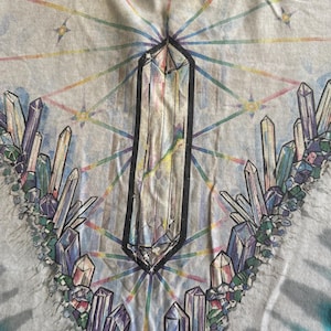 Puede incluir: Una camiseta tie-dye con un dise&ntilde;o de cristal. La camiseta presenta un gran cristal en el centro rodeado de cristales m&aacute;s peque&ntilde;os en varios colores. El fondo es azul claro con un patr&oacute;n tie-dye.