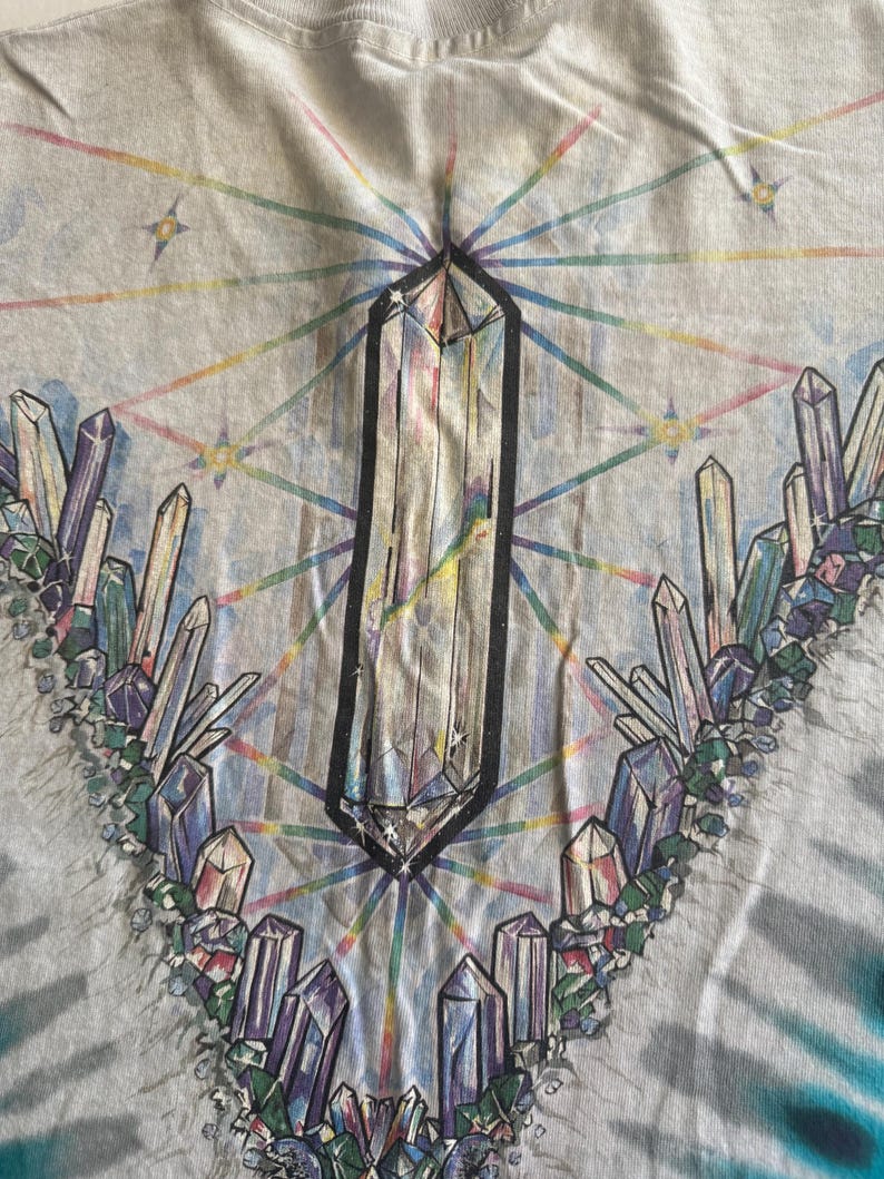 Puede incluir: Una camiseta tie-dye con un dise&ntilde;o de cristal. La camiseta presenta un gran cristal en el centro rodeado de cristales m&aacute;s peque&ntilde;os en varios colores. El fondo es azul claro con un patr&oacute;n tie-dye.