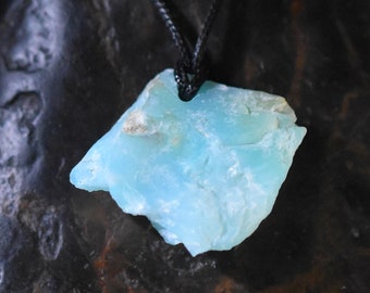 May include: A light blue, raw stone pendant suspended from a black cord. The stone has a rough, natural texture and a pale, sea-foam color. The background is a dark, textured surface.