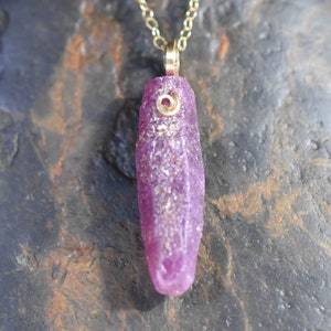 May include: A gold chain necklace with a pendant featuring a raw, elongated, purplish-pink gemstone. The gemstone has a textured surface and a small, gold-colored spiral detail near the top.