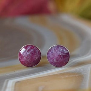 May include: A pair of round stud earrings with a silver setting. Each earring features a faceted, deep pink gemstone. The earrings are displayed on a light-colored, patterned surface.