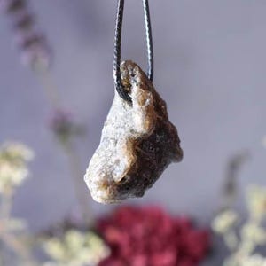 Puede incluir: Un colgante de piedra gris oscuro y marr&oacute;n en un collar de cord&oacute;n negro. La piedra tiene una forma irregular y rugosa y una superficie texturizada.