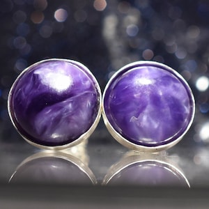 Puede incluir: Un par de pendientes de botón redondos con un bisel plateado. Los pendientes presentan un diseño veteado en morado y blanco. El fondo es un bokeh oscuro y borroso de luces, con los pendientes reflejados en una superficie brillante.