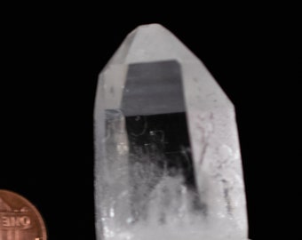 Puede incluir: Una punta de cristal de cuarzo transparente con un interior nublado, descansando sobre una base de piedra gris.
