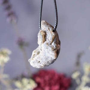 Puede incluir: Un colgante de color gris plateado en forma de media luna con una superficie rugosa y texturizada cuelga de un collar de cord&oacute;n negro.