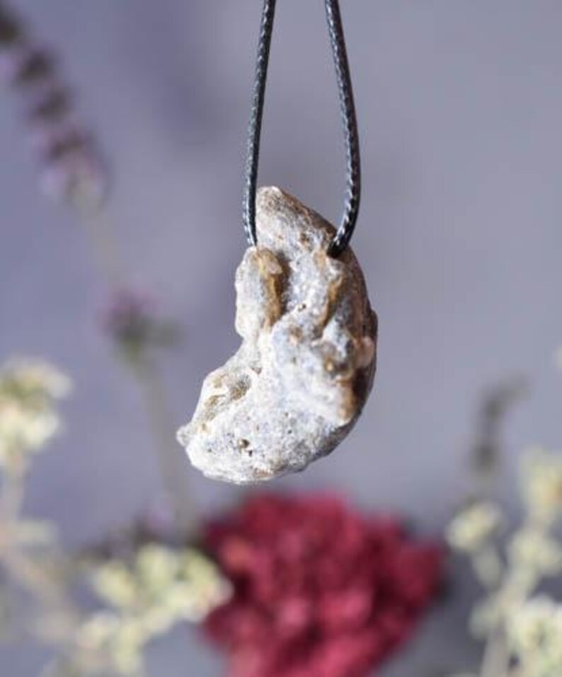 Puede incluir: Un colgante de color gris plateado en forma de media luna con una superficie rugosa y texturizada cuelga de un collar de cord&oacute;n negro.