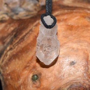 Puede incluir: Un colgante de cristal de cuarzo transparente con un cordón negro. El cristal tiene una forma natural, rugosa y un tono rosa claro.