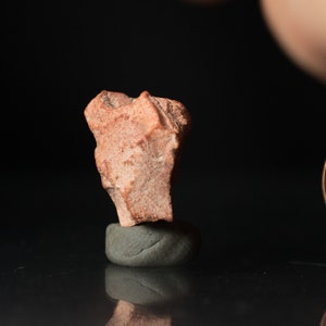 May include: A small, rough-textured, reddish-brown rock fragment is perched atop a smooth, gray, rounded stone. The background is a dark, reflective surface, creating a contrast with the natural tones of the rock.