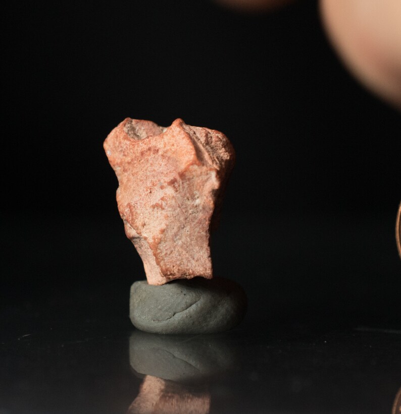 May include: A small, rough-textured, reddish-brown rock fragment is perched atop a smooth, gray, rounded stone. The background is a dark, reflective surface, creating a contrast with the natural tones of the rock.