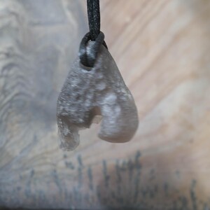 May include: A gray, teardrop-shaped pendant with a rough texture, suspended from a black cord. The pendant has a unique, irregular shape with a cutout section. The background features a blurred, neutral-toned surface.