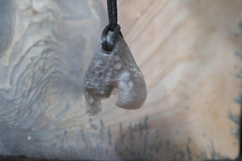 May include: A gray, teardrop-shaped pendant with a rough texture, suspended from a black cord. The pendant has a unique, irregular shape with a cutout section. The background features a blurred, neutral-toned surface.