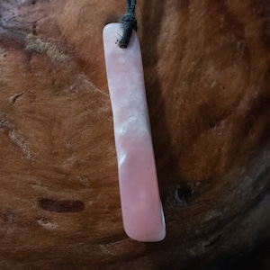 May include: A rectangular pink pendant with a black cord. The pendant has a smooth, polished surface with white marbling. The pendant is suspended against a brown, textured background.