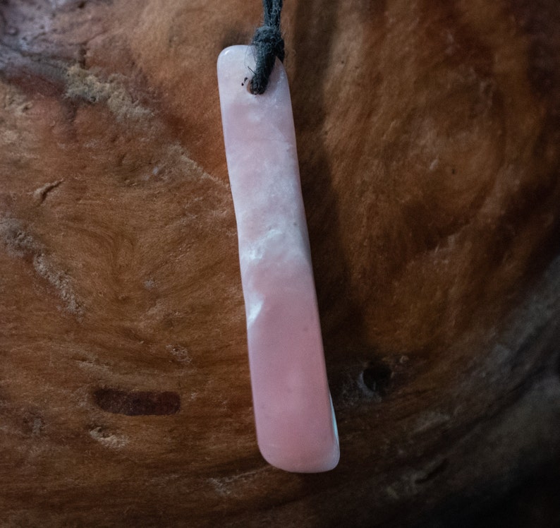 May include: A rectangular pink pendant with a black cord. The pendant has a smooth, polished surface with white marbling. The pendant is suspended against a brown, textured background.