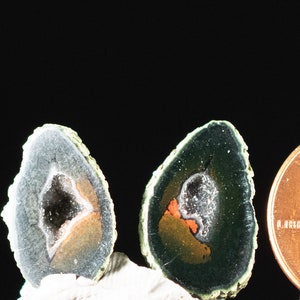 May include: Two sliced geodes with a penny for scale. The geodes have a dark gray exterior with a white, orange, and dark interior. The penny is dated 2014 and shows the profile of Abraham Lincoln.