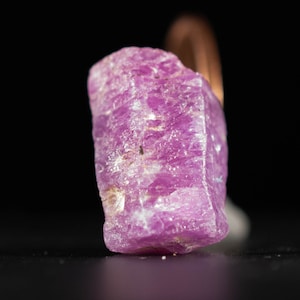 May include: A close-up of a rough, uncut, pink ruby gemstone. The ruby has a crystalline structure and a vibrant, saturated colour. The gemstone is set against a black background.