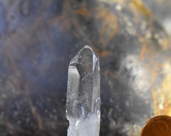 May include: A clear quartz crystal point with a natural, rough texture. The crystal is mounted on a small gray base. The crystal is about 2 inches tall.