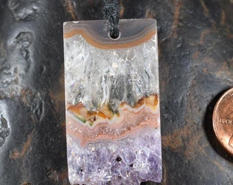 Puede incluir: Colgante rectangular con diseño de piedra natural. El colgante presenta capas de tonos claros, marrones, naranjas, rosas y morados. Una cuerda negra está unida en la parte superior. Una moneda es visible en el fondo.