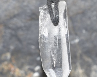 Puede incluir: Un colgante de cristal de cuarzo transparente que cuelga de un cordón negro. El cristal está facetado y tiene una punta puntiaguda. El colgante está colgado sobre un fondo gris.