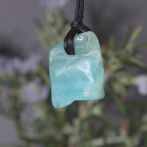 Peut inclure: Pendentif en pierre pr&eacute;cieuse bleu clair avec un cordon noir. La pierre a une surface lisse et polie avec de subtiles variations de couleur. Le pendentif est suspendu &agrave; un cordon noir, cr&eacute;ant un design simple et &eacute;l&eacute;gant. La pierre mesure environ 2.5 cm de long.