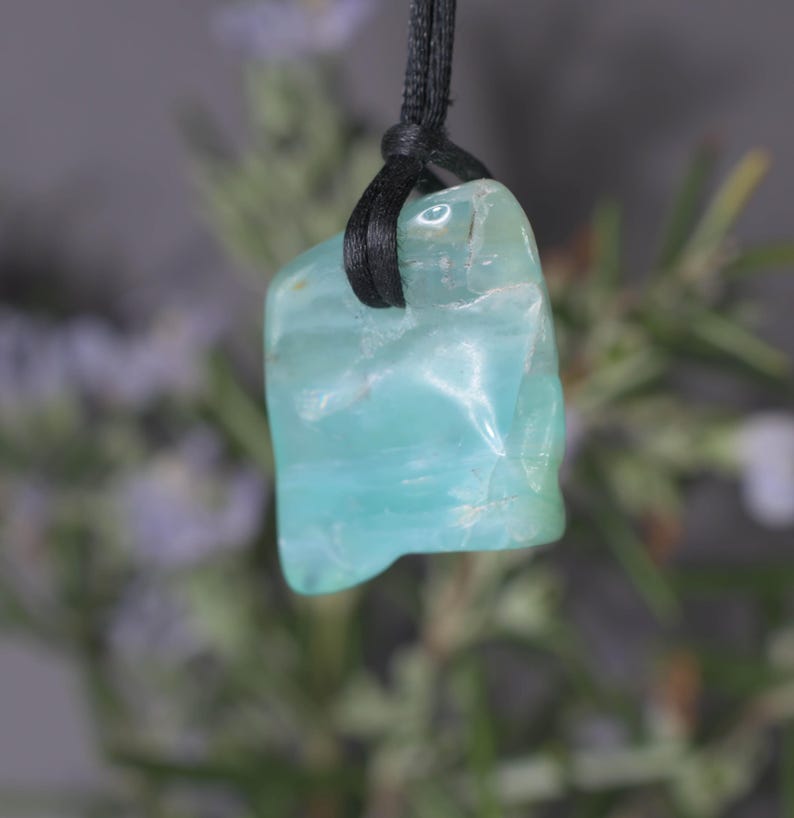 Peut inclure: Pendentif en pierre pr&eacute;cieuse bleu clair avec un cordon noir. La pierre a une surface lisse et polie avec de subtiles variations de couleur. Le pendentif est suspendu &agrave; un cordon noir, cr&eacute;ant un design simple et &eacute;l&eacute;gant. La pierre mesure environ 2.5 cm de long.