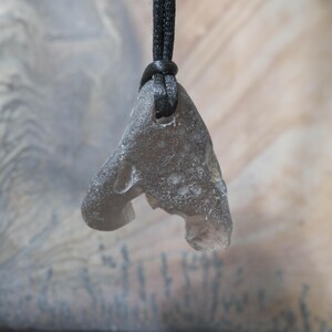 May include: A gray, irregularly shaped pendant made of stone, suspended from a black cord. The pendant has a rough, textured surface and a hole near the top for the cord. The background is a blurred, neutral color.