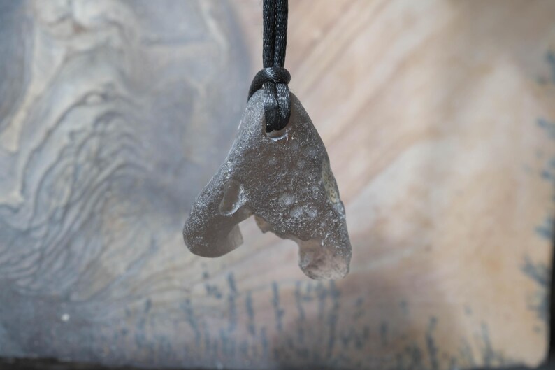 May include: A gray, irregularly shaped pendant made of stone, suspended from a black cord. The pendant has a rough, textured surface and a hole near the top for the cord. The background is a blurred, neutral color.