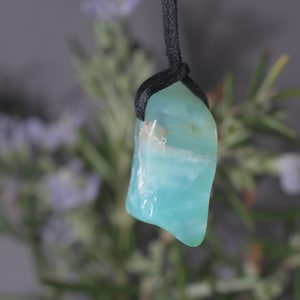 Peut inclure: Un pendentif en pierre polie bleu clair avec un cordon noir. La pierre a une forme lisse et irr&eacute;guli&egrave;re et une qualit&eacute; translucide. L'arri&egrave;re-plan est flou, avec des touches de feuillage vert et violet.