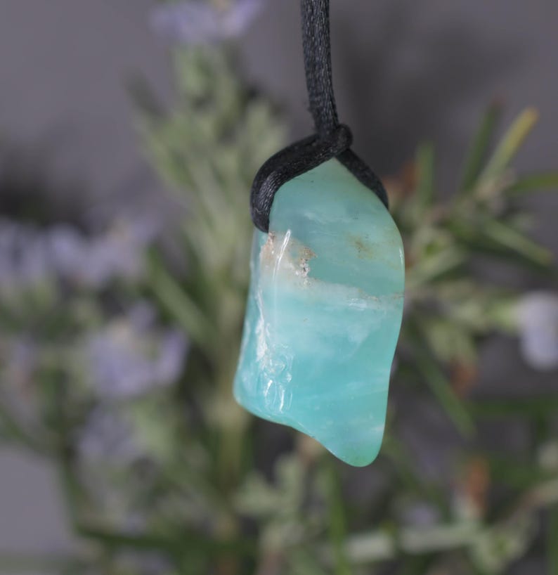 Peut inclure: Un pendentif en pierre polie bleu clair avec un cordon noir. La pierre a une forme lisse et irr&eacute;guli&egrave;re et une qualit&eacute; translucide. L'arri&egrave;re-plan est flou, avec des touches de feuillage vert et violet.
