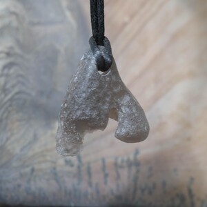 May include: A gray, abstract pendant made of a translucent material, suspended from a black cord. The pendant has an irregular shape with rounded edges and a hole at the top for the cord. The background is a blurred mix of gray and beige.