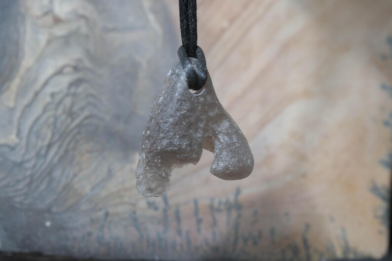 May include: A gray, abstract pendant made of a translucent material, suspended from a black cord. The pendant has an irregular shape with rounded edges and a hole at the top for the cord. The background is a blurred mix of gray and beige.